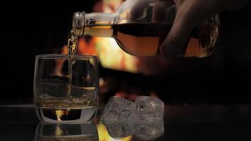 Pouring whiskey or cognac from a Bottle to a Glass over Fireplace background. Warmth and home comfort. 4K video