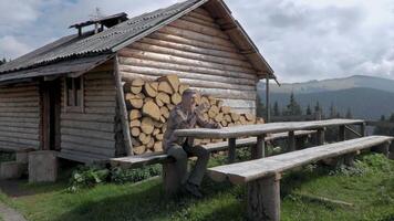 un hombre es descansando cerca un antiguo de madera casa en el montañas. el real hogar de cárpato pastores en el montañas. concepto de aventura, viaje y escapar desde el ciudad. lento movimiento imágenes 4k video