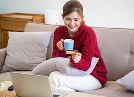 un joven asiático mujer con un contento sonrisa sostiene un crédito tarjeta y usos un teléfono inteligente a tienda en línea en línea pago concepto. foto