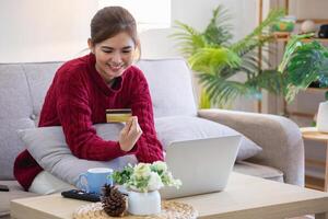 un joven asiático mujer con un contento sonrisa sostiene un crédito tarjeta y usos un teléfono inteligente a tienda en línea en línea pago concepto. foto