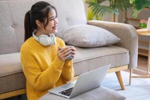 hermosa joven mujer en un amarillo casual vestir disfrutando escuchando a música y sonriente mientras relajante en el sofá a hogar. joven mujer con auriculares usos ordenador portátil y teléfono inteligente a hogar. relajarse concepto foto