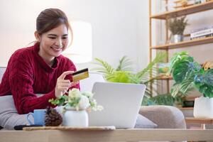 un joven asiático mujer con un contento sonrisa sostiene un crédito tarjeta y usos un teléfono inteligente a tienda en línea en línea pago concepto. foto