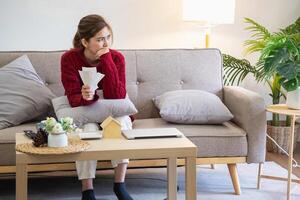 A young Asian woman sits on a sofa in her home, feeling worried and frustrated about her monthly expenses. Various utility bills. photo