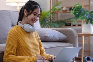 Beautiful young woman in a yellow casual dress enjoying listening to music and smiling while relaxing on the sofa at home. Young woman with headphones uses laptop and smartphone at home. relax concept photo