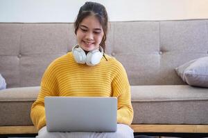 Beautiful young woman in a yellow casual dress enjoying listening to music and smiling while relaxing on the sofa at home. Young woman with headphones uses laptop and smartphone at home. relax concept photo