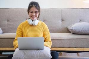 Beautiful young woman in a yellow casual dress enjoying listening to music and smiling while relaxing on the sofa at home. Young woman with headphones uses laptop and smartphone at home. relax concept photo