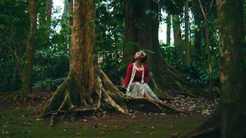 Woman in red top sitting in a lush green forest, looking upwards with a sense of wonder. video