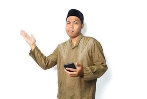 confused asian moslem man wearing islamic clothes presenting to beside with holding mobile phone isolated on white background photo