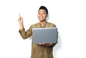 smiling asian muslim man holding laptop pointing to above isolated on white background photo