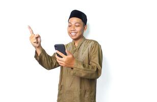 excited asian muslim male holding mobile phone and pointing to beside isolated on white background photo