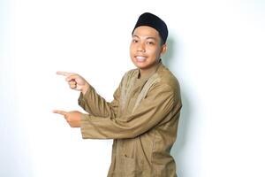 asian muslim man pointing to the right side and smiling at camera wearing islamic dress isolated on white background photo