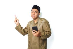 confused asian muslim man wearing islamic clothes pointing to above with holding mobile phone isolated on white background photo