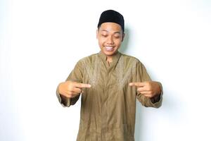 smiling asian muslim man wearing koko clothes pointing to the middle with excitement expression looking at camera isolated on white background photo