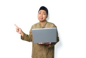 crying asian muslim man holding laptop and pointing to beside isolated on white background photo