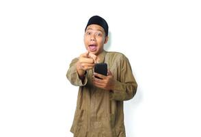 surprised asian muslim man wearing islamic clothes pointing at camera with holding mobile phone isolated on whtie background photo