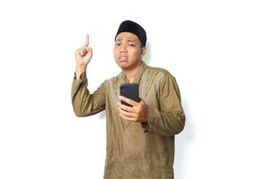 anxious asian muslim man holding mobile phone with pointing to above show worry expression isolated on white background photo