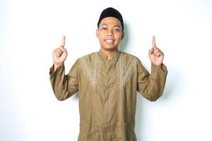 asiático musulmán hombre señalando a encima y sonriente a cámara vistiendo islámico vestir aislado en blanco antecedentes foto