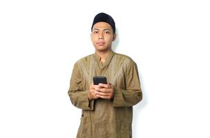 confused asian muslim man wearing islamic clothes using smartphone, looking at camera, isolated on white background photo