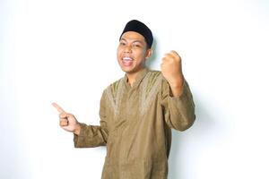 joyful asian muslim man pointing to empty space and raising fist show winner gesture in eid mubarak celebration isolated in white background photo