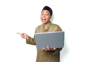 surprised asian muslim man pointing to beside with holding laptop isolated on white background photo