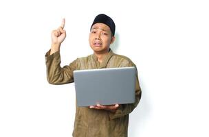 crying asian muslim man holding laptop and pointing above isolated on white background photo