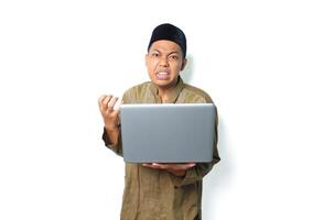 asian muslim man holding laptop with angry expression wearing islamic dress isolated on white background photo