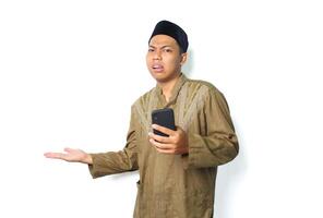 confuso asiático musulmán hombre vistiendo islámico ropa presentación a junto a con participación móvil teléfono aislado en blanco antecedentes foto