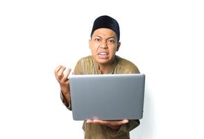 asian muslim man holding laptop with angry expression wearing islamic dress isolated on white background photo