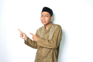 confused asian muslim man pointing to beside and looking at camera wearing koko clothes isolated on white background photo