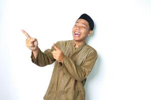 ecstatic asian muslim man wearing islamic dress pointing to empty side with laugh isolated on white background photo