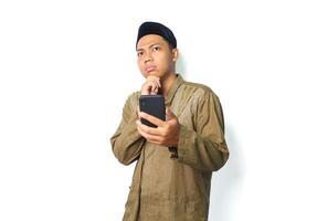 thoughtful asian muslim man holding mobile phone with thouching his chin looking away isolated on white background photo