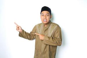 angry asian muslim man wearing arabic costume pointing to side and look at camera isolated on white background photo