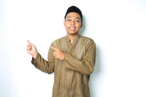 asian muslim man pointing to the right side and smiling at camera wearing islamic dress isolated on white background photo
