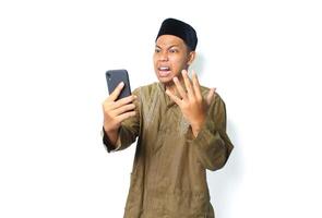 enojado asiático musulmán hombre vistiendo islámico vestir participación móvil teléfono mirando a cámara aislado en blanco antecedentes foto