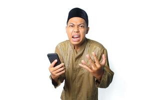 enojado asiático musulmán hombre vistiendo islámico vestir participación móvil teléfono mirando a cámara aislado en blanco antecedentes foto
