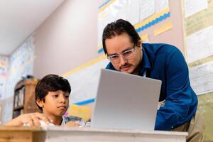 Diverse male teacher and schoolboy using laptop in elementary school class photo