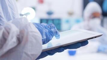 Close up handheld footage of chemist typing on tablet in a research laboratory. Team of scientist working in the background. Scientist in coverall equipment. video