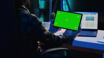 Man in home office checking statistical financial graphs on laptop, crosschecking with data on mockup tablet. Remote employee holding green screen device, analyzing business graphics video