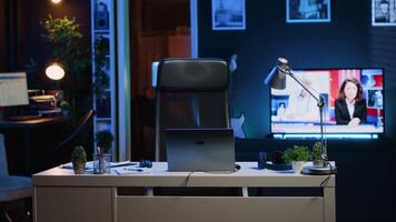 Office desk filled with business productivity items in empty home interior with TV left open in background at night. Laptop, headphones, plants and work documents on table in apartment room, camera A video