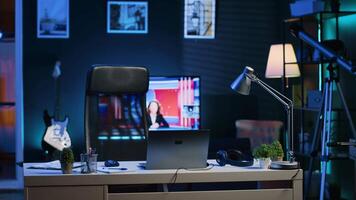 Focus on office desk in front of smart TV in blurry background displaying news program broadcasting. Laptop on table and television screen in empty room showing VOD channel, camera A video