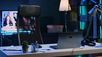 Dimly lit apartment with office desk in front of smart TV displaying news program broadcasting. Laptop on table and television screen in empty room showing channel, camera A close up shot video