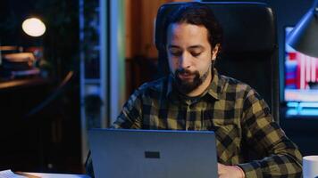 sorridente homem sentado às casa escritório escrivaninha e digitando em computador portátil teclado, resolução tarefas. feliz controlo remoto trabalhador usando caderno para mandar e-mails para o negócio parceiros a partir de vivo sala, Câmera uma video