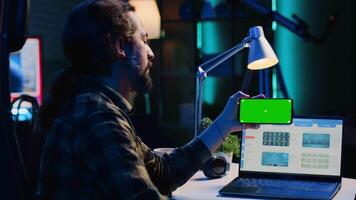 Teleworker in apartment office checks statistics on laptop, crosschecking with data on green screen cellphone. Person holding mockup mobile phone in landscape mode, analyzing company revenue video