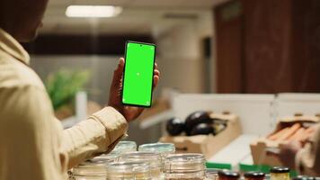 African american storekeeper holding phone with greenscreen in local grocery store, having organic products on sale. Vendor presents isolated chromakey display on smartphone, mockup layout. Camera A. video