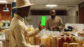 affaires propriétaire en présentant Vide écran vert afficher sur téléphone, montrant isolé maquette disposition dans zéro déchets éco magasin. africain américain vendeur en utilisant chromakey modèle sur mobile téléphone. caméra un. video