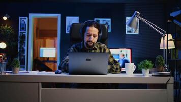 Caucasian man sitting on chair at home office desk in neon illuminated apartment, working and listening music. Teleworker wearing headphones in RGB lit living room, doing his job remotely video