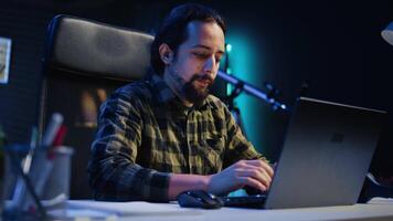 Freelancer sitting at home office desk and typing on laptop keyboard, doing his job. Remote worker using electronic device to send emails to customers from living room, camera B video