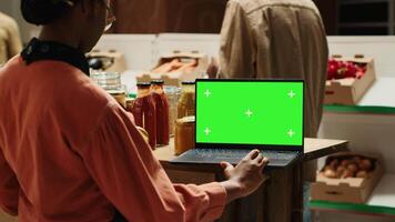 Young vendor looks at greenscreen on laptop while preparing merchandise for customers, using isolated mockup layout on screen. African american seller checking blank chromakey display. Camera A. video