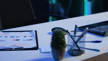 Office desk filled with productivity items in empty home interior illuminated with neon lights at night. Laptop on table and other work related objects in apartment room, camera A close up shot video