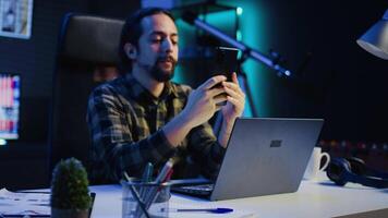 Man relaxing at home sitting at office desk, texting friends on smartphone in front of laptop. Cheerful freelancer taking break to chat with mates online using mobile phone in living room, camera B video